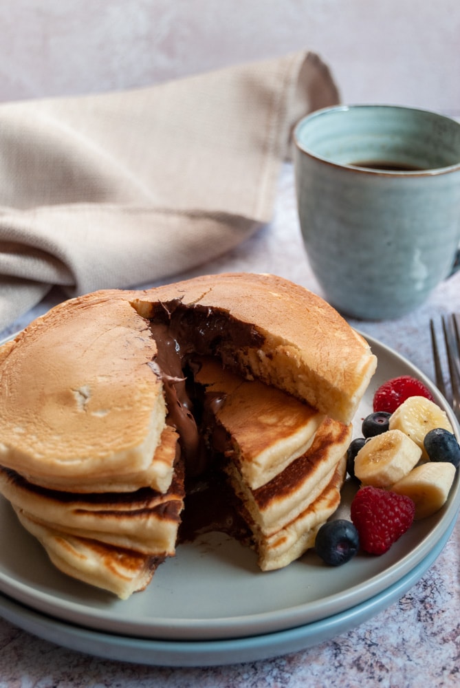 a stack of pancakes filled with Nutella chocolate spread with fresh sliced bananas, blueberries and raspberries on a light grey plate, a blue cup of coffee and a beige linen napkin on a marbled grey worktop.