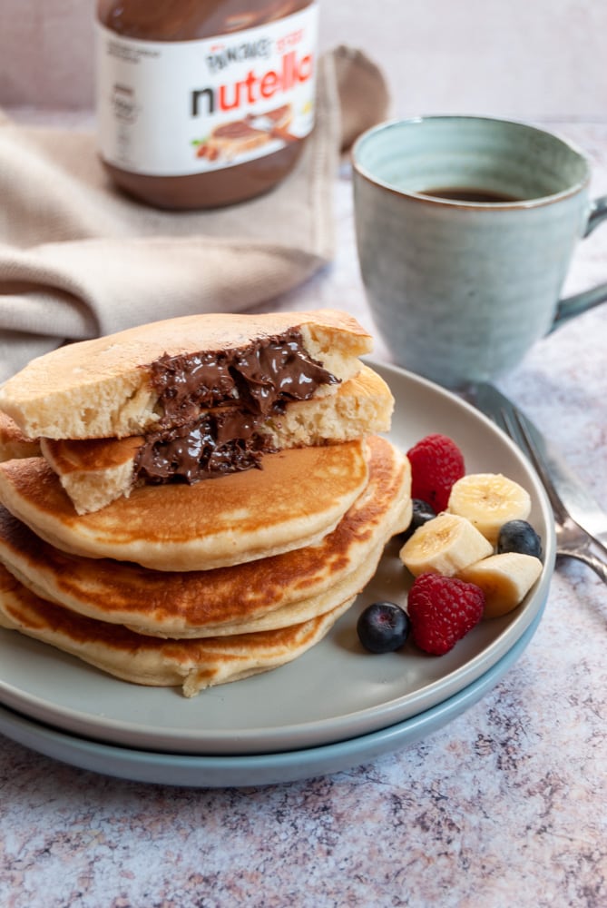 a stack of four pancakes on a light grey plate with fresh sliced bananas, blueberries and raspberries. The top pancake has been broken in half to reveal an melted Nutella filling. A blue cup of coffee, a beige linen napkin and a jar of Nutella sits in the background.