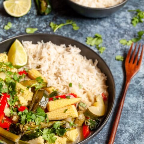 a black stoneware bowl of vegetable curry with white rice sprinkled with coriander, a wooden fork, a small black bowl of rice with a wooden spoon and a lime wedge on a dark blue background.