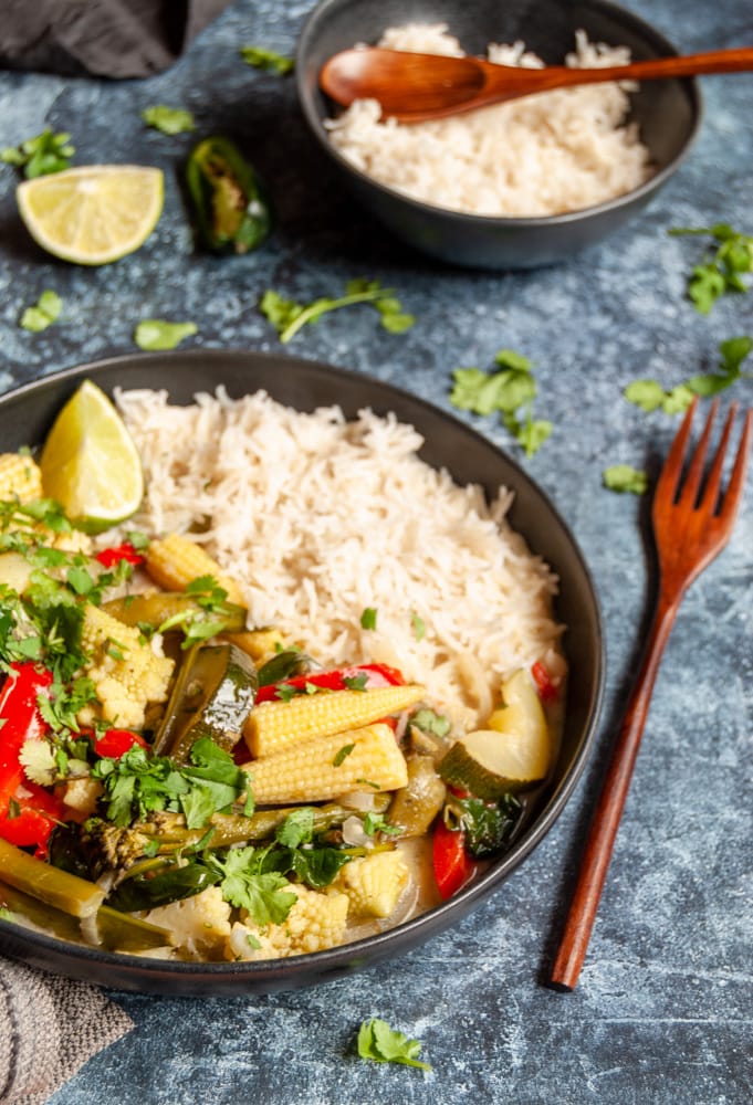 a black stone ware bowl of green Thai vegetable curry with baby corn, courgettes, red pepper and broccoli with white rice on a dark blue background, a wooden fork, and a small black bowl of rice.  Fresh coriander leaves are scattered around the bowl of curry.