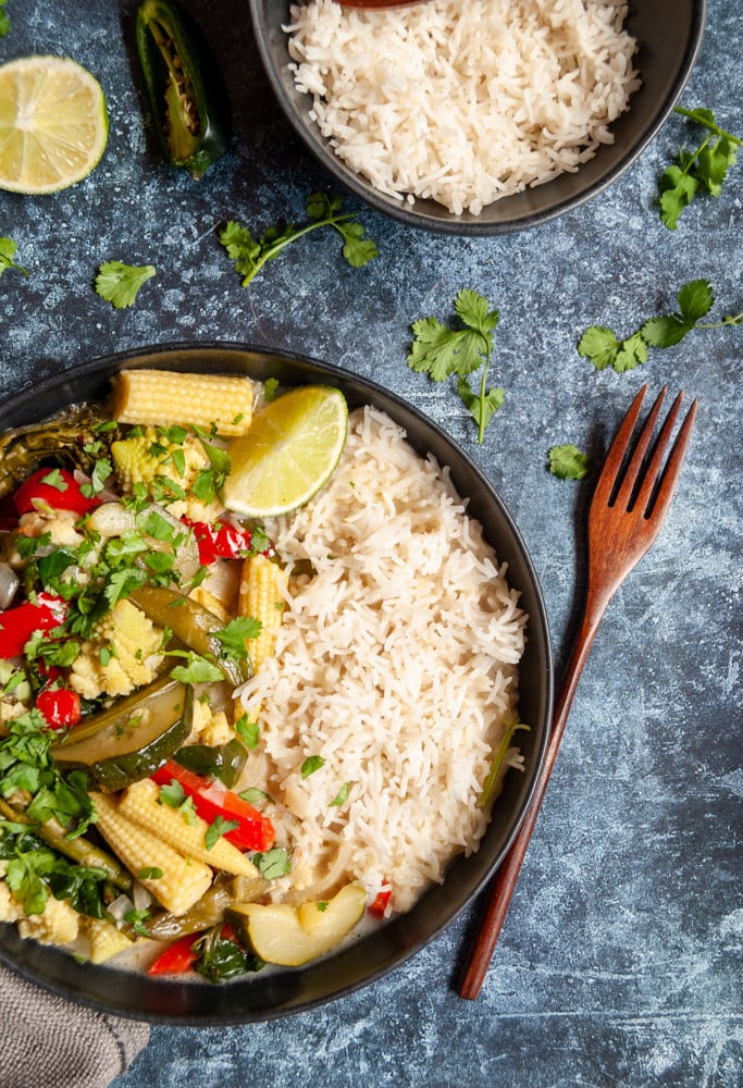 a bowl of Thai Green vegetable curry sprinkled with coriander and white rice, a wooden fork, a small black bowl of rice and a lime wedge on a dark blue backdrop.  Fresh coriander leaves are scattered around the plates.