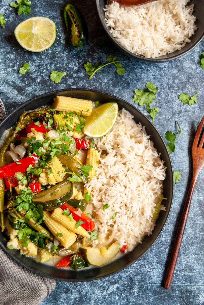 a black stoneware bowl of Thai curry with red peppers, baby corn and courgettes sprinkled with fresh coriander, a wooden fork, a small bowl of rice and a lime wedge on a dark blue background.  Fresh coriander stalks are scattered around the dishes