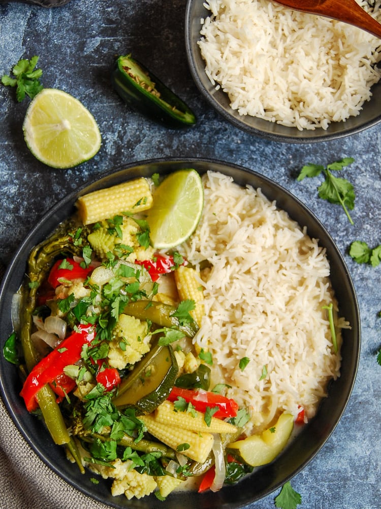 a black stoneware bowl of Thai Green curry with red peppers, baby corn, zucchini, and broccoli with white rice and a lime wedge on a dark blue and white backdrop.  A bowl of rice can be partially seen in the top right corner.  Sprigs of fresh coriander and a piece of lime are sitting beside the bowl of curry.