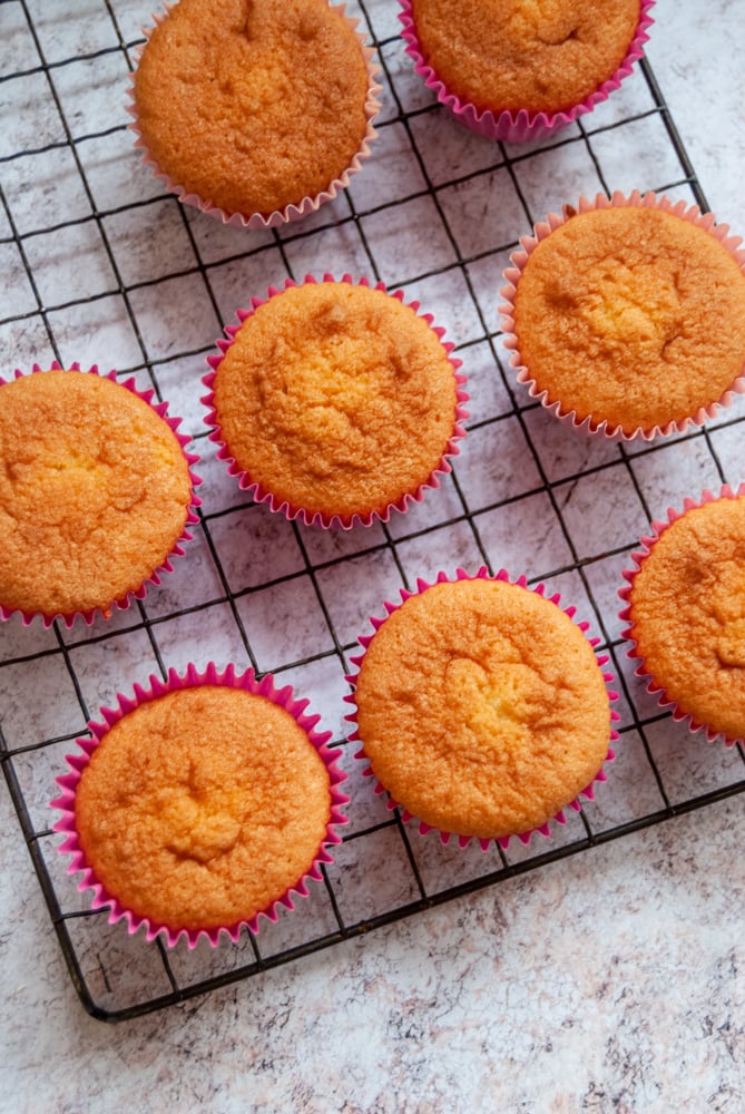 eight vanilla cupcakes in pink wrappers on a black wire rack and marbled grey worktop.