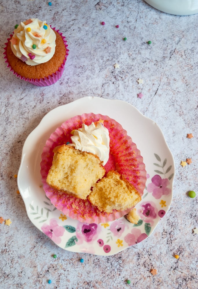 a cupcake topped with vanilla buttercream broken in half, a pink cupcake wrapper on a pink floral plate.