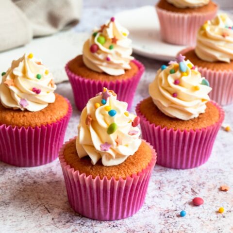 A batch of vanilla cupcakes topped with buttercream frosting and coloured sprinkles. A cupcake on a plate can be seen in the background. Sprinkles are scattered all around the cupcakes.