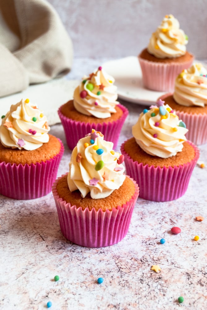 A batch of vanilla cupcakes topped with buttercream frosting and coloured sprinkles. A cupcake on a plate can be seen in the background. Sprinkles are scattered all around the cupcakes.