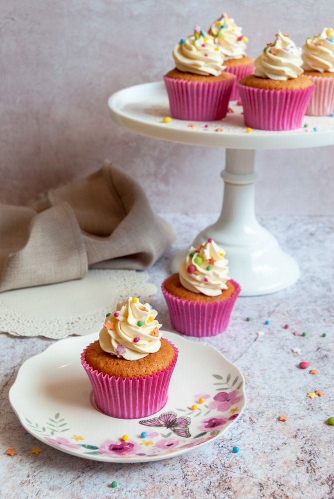 A cupcake in a pink cupcake wrapper topped with vanilla buttercream and coloured sprinkles on a pink floral plate and more cupcakes on a white cake stand. Sprinkles are scattered all around the cakes. A paper doily and a beige napkin are sitting beside the cake stand.