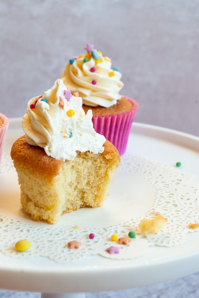 a vanilla cupcake topped with vanilla buttercream frosting and coloured sprinkles on a white cake stand.