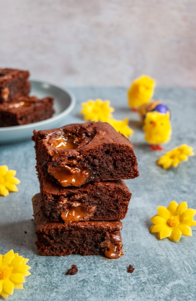 a stack of three caramel egg filled chocolate brownies on a light blue and white backdrop. Yellow flowers are sitting beside the brownies and a blue plate of brownies and Easter chicken toys can be partially seen in the background.