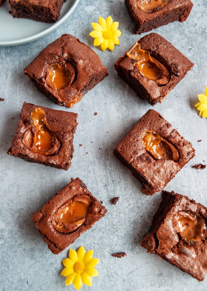 a flat lay photo of chocolate brownies topped with chocolate eggs with a molten caramel filling and yellow flowerson a blue backdrop.