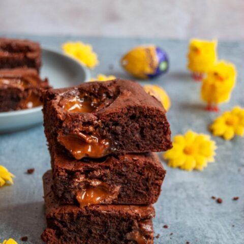 a stack of three chocolate brownies filled and topped with caramel filled easter eggs on a light blue backdrop. Yellow flowers, Easter chicken toys and a blue plate of the brownies can be partially seen in the background.