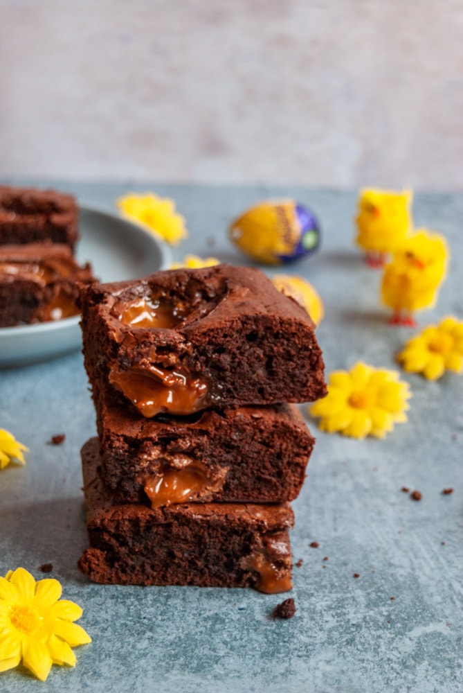 a stack of three chocolate brownies filled and topped with caramel filled easter eggs on a light blue backdrop. Yellow flowers, Easter chicken toys and a blue plate of the brownies can be partially seen in the background.