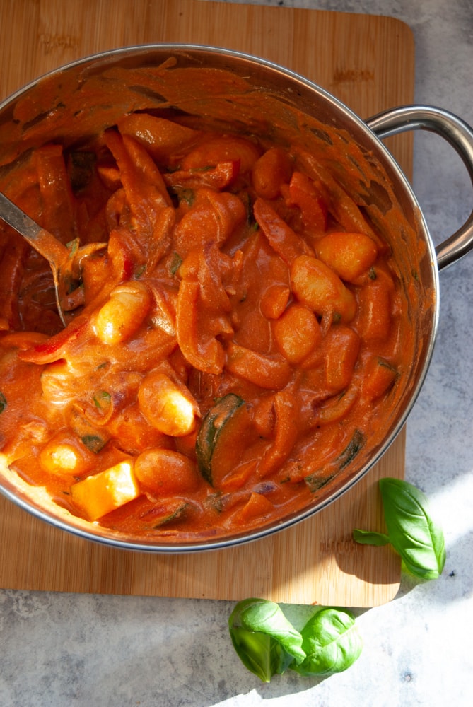 an overhead image of a silver pan of gnocchi in a tomato mascarpone sauce on a wooden board with fresh basil leaves.