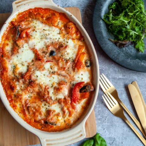 an overhead image of an oval casserole dish of baked gnocchi in a tomato sauce with peppers and olives. A black rustic plate with green salad and gold knives and forks are sitting beside the casserole.