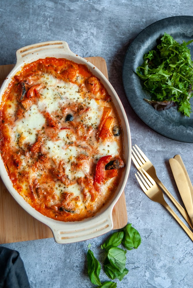 an overhead image of an oval casserole dish of baked gnocchi in a tomato sauce with peppers and olives.  A black rustic plate with green salad and gold knives and forks are sitting beside the casserole.