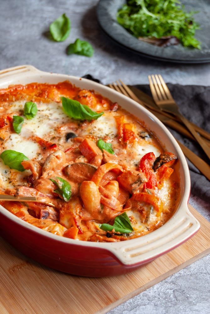 A gnocchi bake with tomato sauce, peppers and olives in a red and white oval casserole dish on a wooden board, gold cutlery on a black napkin and a black plate of green salad.