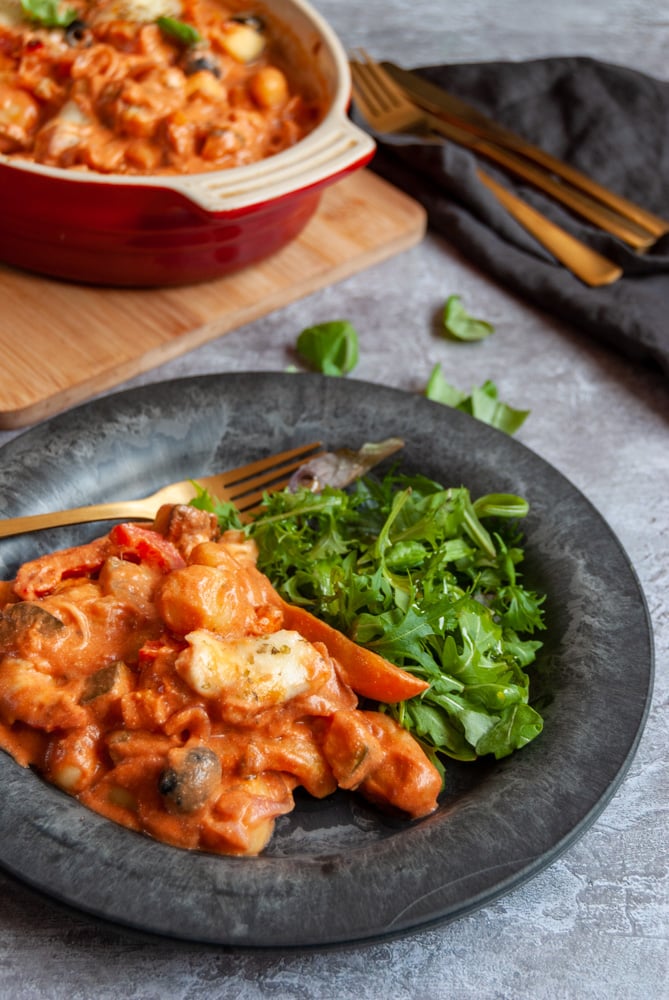 a black plate of a creamy gnocchi bake with tomato sauce, onions, peppers and olives with a green salad. Gold cutlery on a black napkin and a red and white casserole dish of the gnocchi bake can be seen in the background.