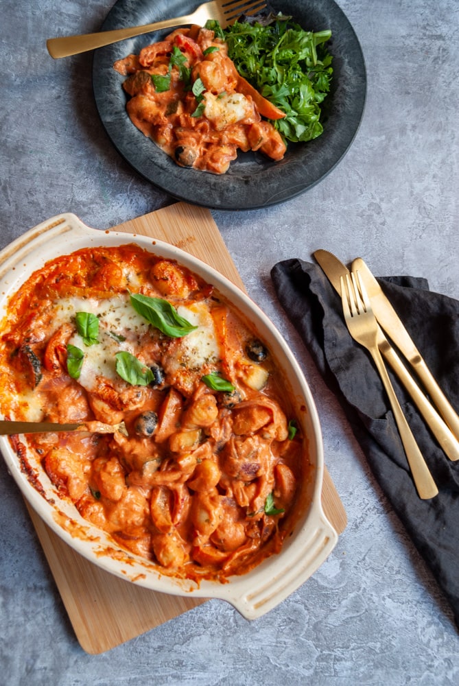 an overhead shot of a white oval casserole dish of gnocchi, peppers, onions and olives in a creamy tomato sauce with mozzarella, gold cutlery on a black napkin and a black plate of the gnocchi bake with a green salad.