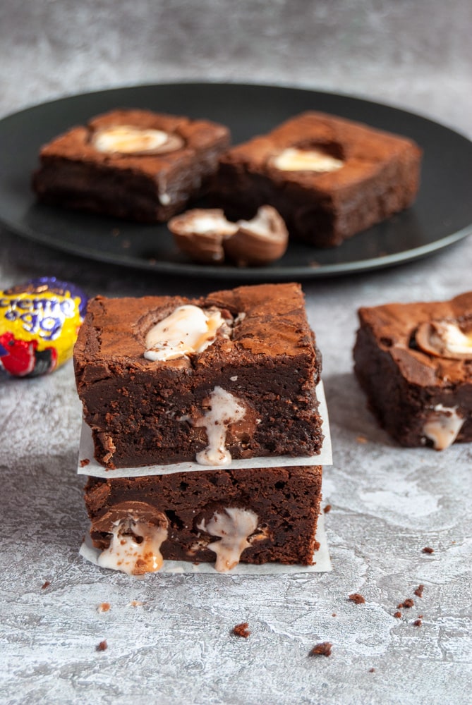 two chocolate brownies filled and topped with creme eggs stacked on top of each other on a grey backdrop.  More brownies on a black plate in the background.