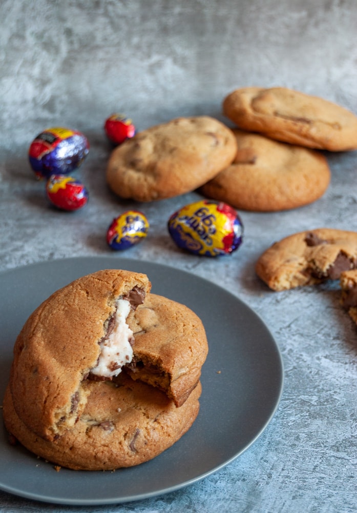 two large chocolate chunk cookies on a grey plate. One of the cookies has been broken in half to reveal a creme egg middle. More cookies and creme eggs can be seen in the background.