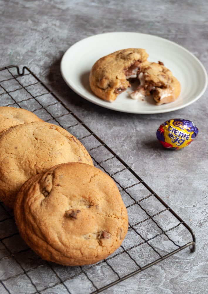 chocolate chunk cookies on a black wire rack/grey backdrop.  A white plate with a cookie broken in half to reveal a creme egg filling.