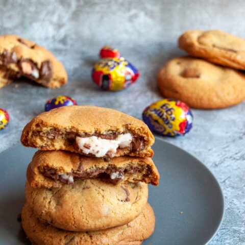 three large chocolate chunk cookies on a grey plate. The top cookie has been broken in half to reveal a molten creme egg middle. More cookies and creme eggs can be seen in the background.