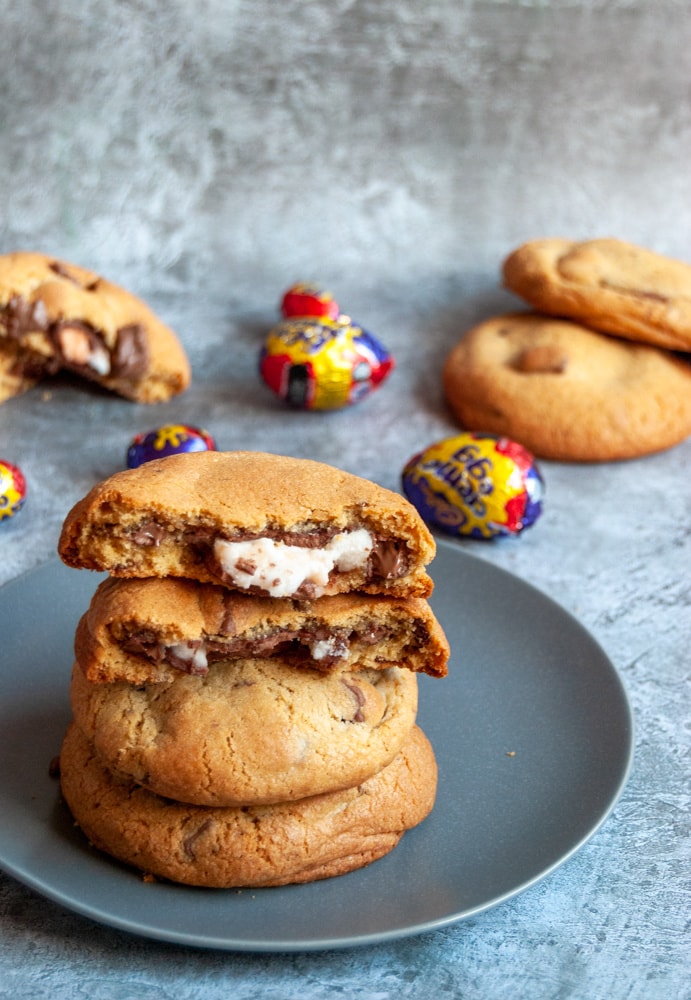 three large chocolate chunk cookies on a grey plate.  The top cookie has been broken in half to reveal a molten creme egg middle. More cookies and creme eggs can be seen in the background.