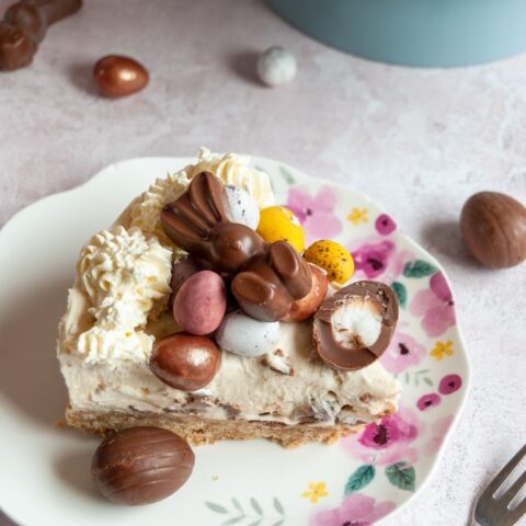 a slice of cheesecake topped with whipped cream and Easter chocolate on a floral plate with a mini chocolate egg, a small cake fork and mini Easter egg chocolates.