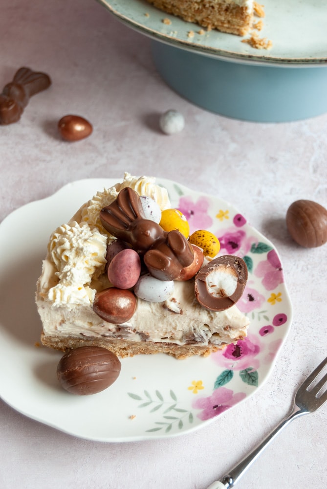 a slice of cheesecake topped with whipped cream and Easter chocolate on a floral plate with a mini chocolate egg, a small cake fork and mini Easter egg chocolates.