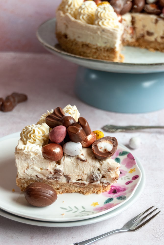 a slice of cheesecake topped with whipped cream and chocolates on a floral plate.  A larger cheesecake on a blue plate can be partially seen in the background.