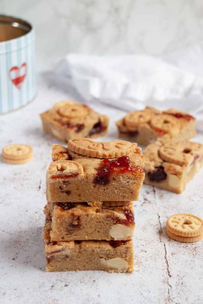 a stack of three blondies filled with white chocolate chips, strawberry jam swirls and topped with a jammie dodger biscuit on a white and grey backdrop.  More blondies, a blue and red cake tin and a white napkin are in the background.