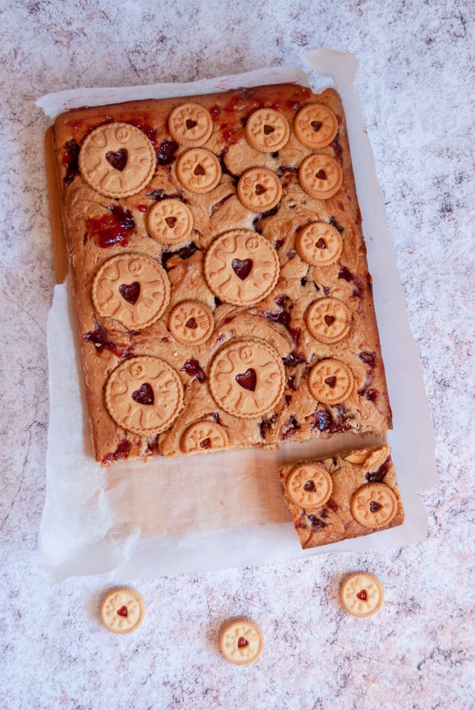 a partially sliced blondie bar with jam swirls and jammie dodger biscuits on a piece of baking parchment.