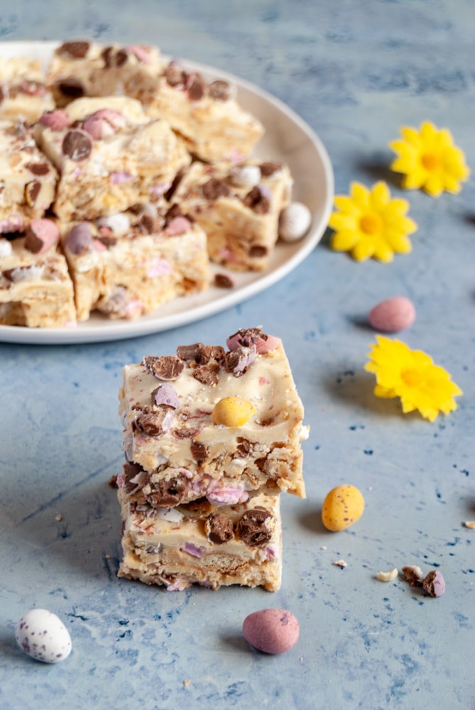 two pieces of white chocolate rocky road topped with mini chocolate Easter eggs sitting on top of each other. A white plate with more rocky road squares can be seen in the background along with artificial yellow flowers and mini coloured chocolate eggs.