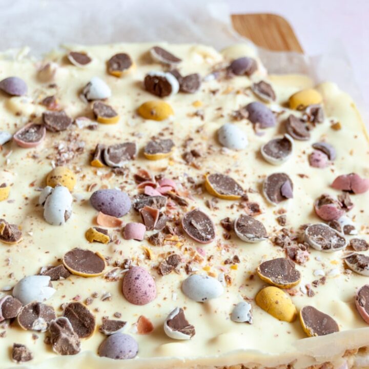 a large piece of unsliced rocky road topped with white chocolate and mini coloured Easter egg chocolates on a wooden board.