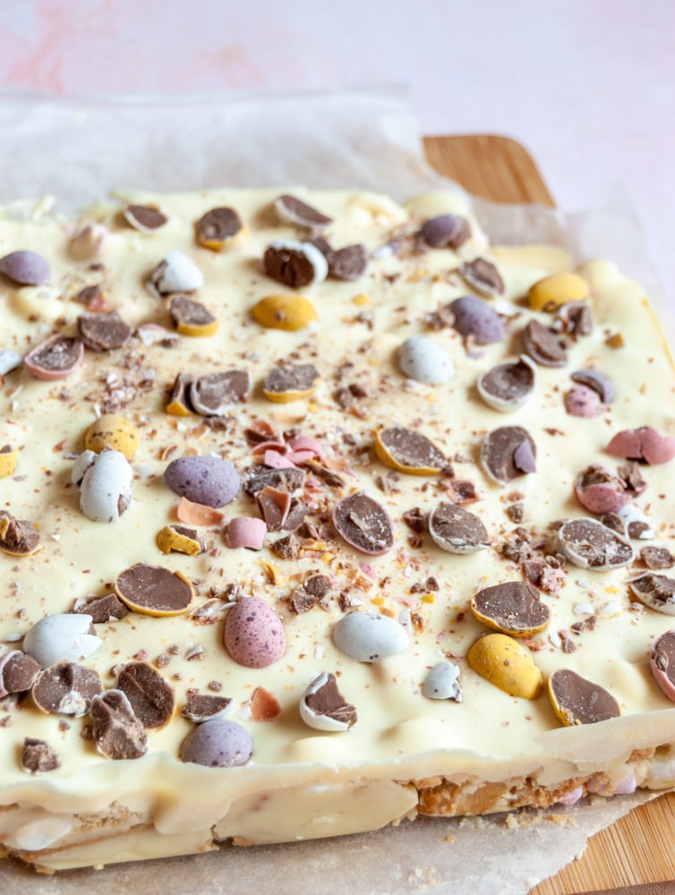 a large piece of unsliced rocky road topped with white chocolate and mini coloured Easter egg chocolates on a wooden board.