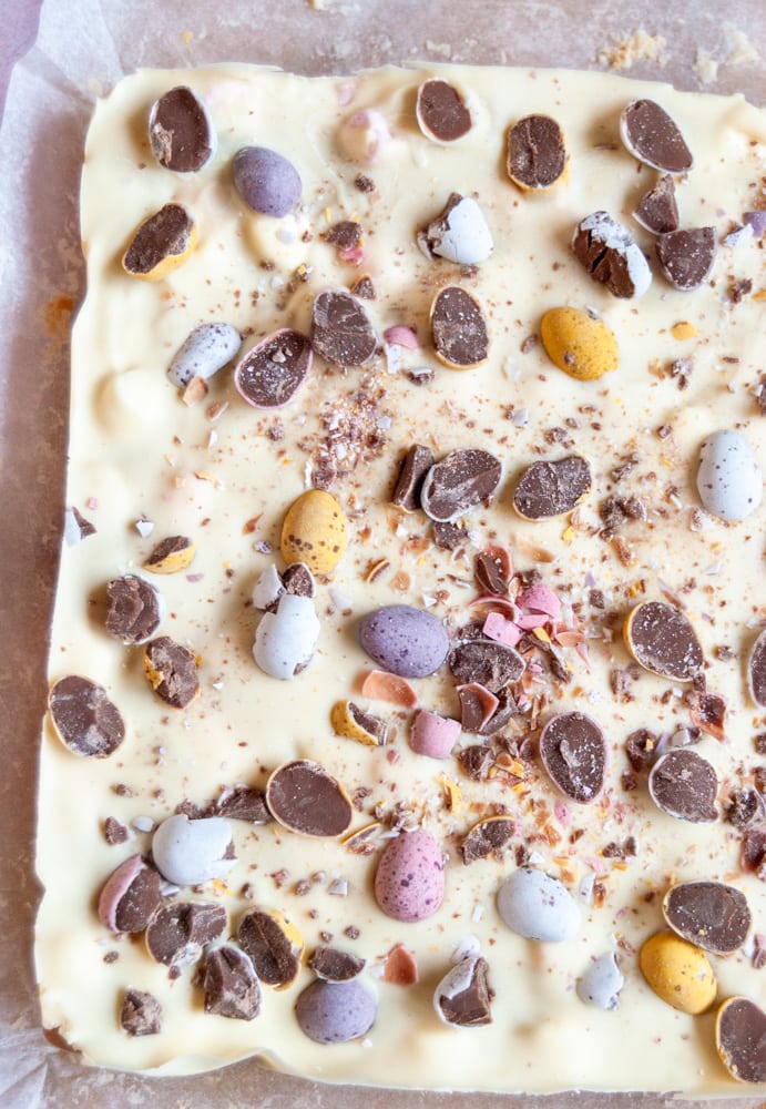 an overhead image of a large unsliced piece of rocky road topped with melted white chocolate and coloured mini Easter egg chocolates.