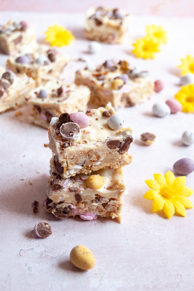 squares of white chocolate rocky road with marshmallows and mini chocolate eggs on a light grey and pink backdrop with mini chocolate eggs and small yellow flowers.