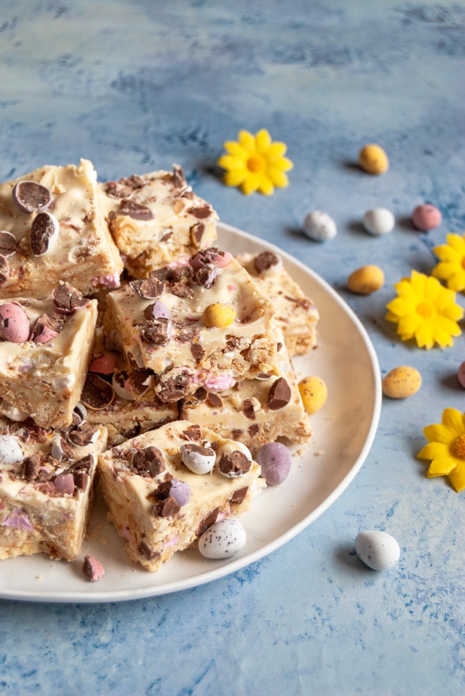 a white plate of white chocolate rocky road topped with mini coloured chocolate eggs on a blue backdrop. more chocolate eggs and yellow flowers are placed around the plate.