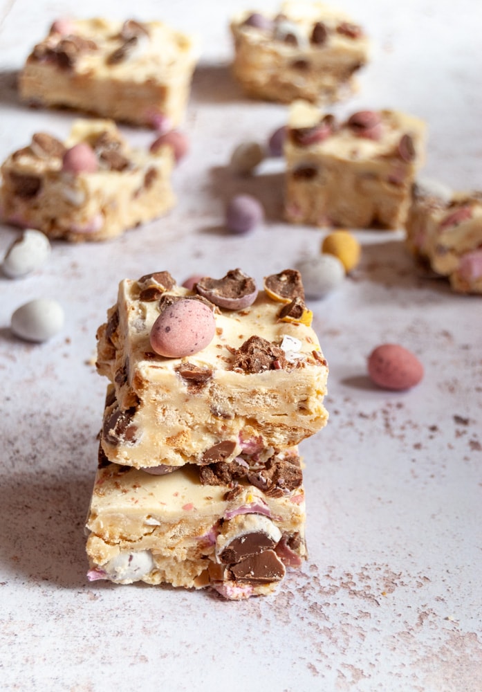 a close up image of two pieces of white chocolate rocky road topped with mini pink and purple rocky road. more rocky road pieces can be seen in the background.