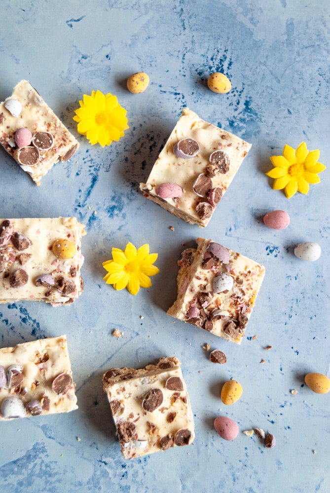 a flat lay image of rocky road squares topped with mini coloured chocolate eggs, little yellow artificial flowers and more mini eggs on a duck egg blue backdrop.