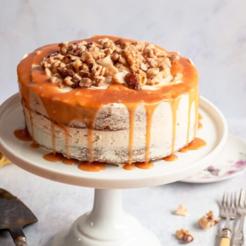 A banana cake topped with chopped nuts and salted caramel glaze on a white cake stand.