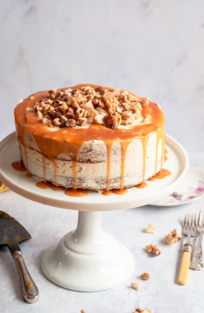 A banana cake topped with chopped nuts and salted caramel glaze on a white cake stand.