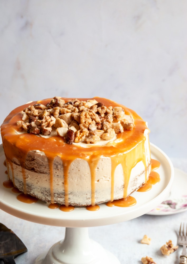 a banana cake covered in caramel frosting, salted caramel sauce and chopped nuts on a white cake stand.  The backdrop is a greyish white.