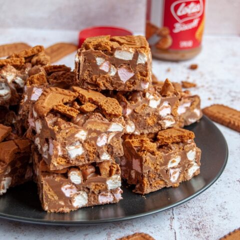 A black plate with squares of rocky road with Biscoff cookies. A jar of biscoff spread can be seen in the background.