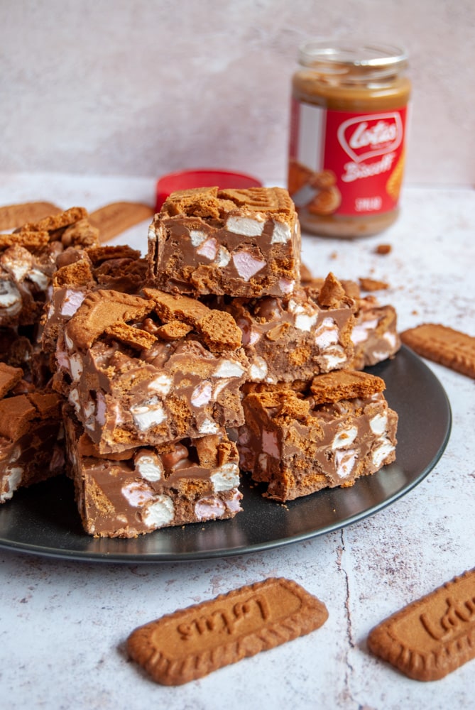 A black plate with squares of rocky road with Biscoff cookies. A jar of biscoff spread can be seen in the background.