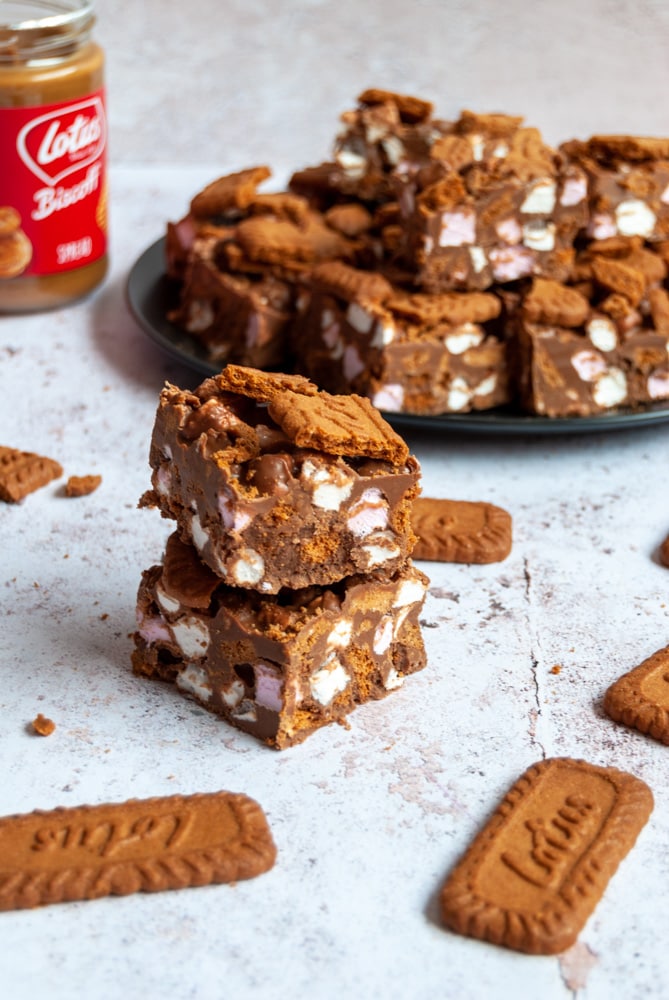 two pieces of rocky road topped with Biscoff cookies on a white distressed backdrop, a black plate of the rocky road squares piled on top of each other and a jar of biscoff spread and biscoff cookies.