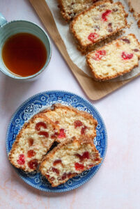 three slices of cherry loaf cake on a blue willow pattern plate, a blue stoneware mug of tea and more slices of the cake on a wooden board