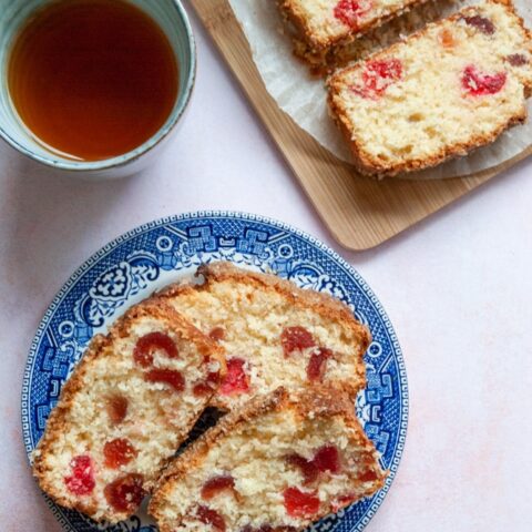 three slices of cherry loaf cake on a blue willow pattern plate, a blue stoneware mug of tea and more slices of the cake on a wooden board