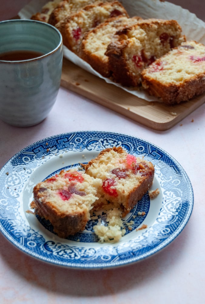 A slice of crumbly cherry cake on a blue willow pattern plate, a stoneware blue mug of tea and more slices of cherry cake on a wooden board.